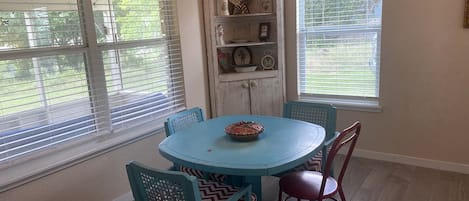 Un coin repas comprenant une table ronde, quatre chaises, une étagère décorative et de grandes fenêtres équipées de stores.