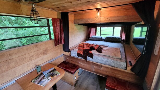 Interior acolhedor de cabana com teto de madeira, cama com cobertor, mesa pequena com revista e janela com cortinas.