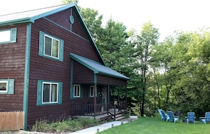 A two-story wooden house with a porch and a deck, surrounded by a grassy yard and trees. - Lakefront Vacation Rental with a Deck and Hot Tub near Wisconsin Dells (Tomah)