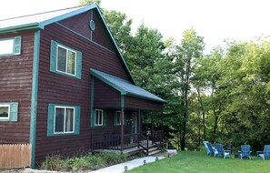 Ein zweistöckiges Holzhaus mit Veranda und Terrasse, umgeben von einem Rasen und Bäumen.