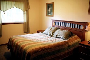 Ein Schlafzimmer mit Bett, zwei Kissen, einem Nachttisch, einer Lampe, einem Fenster mit Vorhängen und einem gerahmten Bild an der Wand.