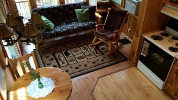 A cozy cabin interior with a wooden dining table, a rocking chair, a sofa with patterned cushions, and a kitchen area with a stove.
