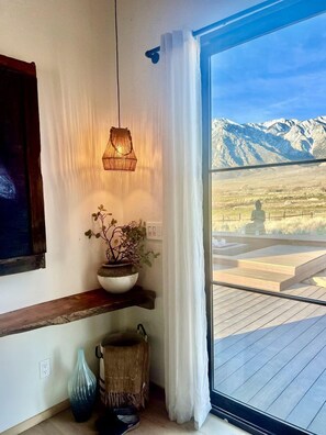 Una habitació amb una finestra gran, un llum penjant, una planta en test, una prestatgeria de fusta i vistes a les muntanyes.