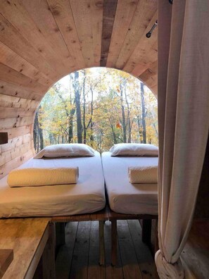 A cabin with two beds, a wooden ceiling, and a circular window offering a view of autumn trees.
