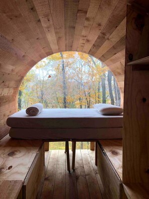 A wooden cabin with a circular skylight, a bed with pillows, and a view of autumn trees.