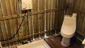 Een buitentoilet met houten omheining, voorzien van een wit toilet, een klein wastafeltje en een verwarming.