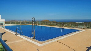 A rectangular outdoor swimming pool with a metal ladder, surrounded by a tiled deck, and overlooking a landscape with hills and a clear sky.