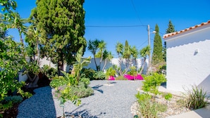 Un jardín con un sendero de grava, diversas plantas y un edificio blanco con techo de tejas.