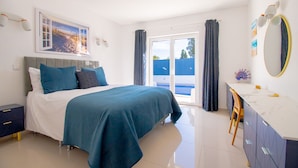 Un dormitorio con cama, escritorio, silla, ventana grande y un cuadro en la pared.