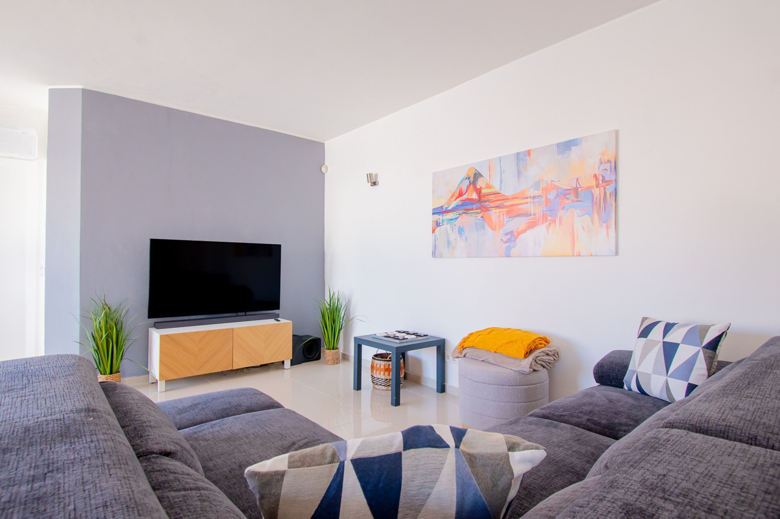 A modern living room with a flat-screen TV, grey sofas, a coffee table, and a colorful abstract painting on the wall.
