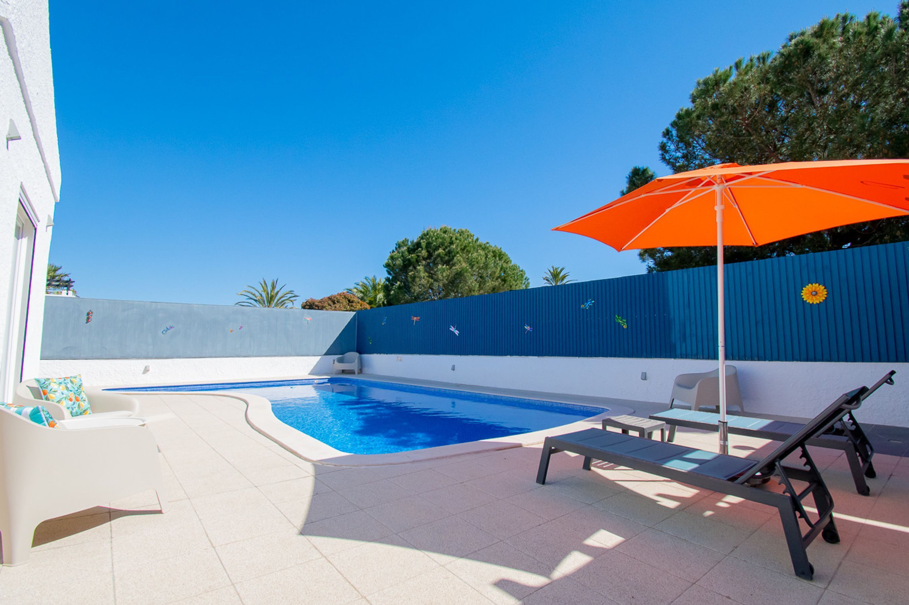 Piscina con sillas de descanso y una sombrilla, rodeada por un muro azul y árboles.
