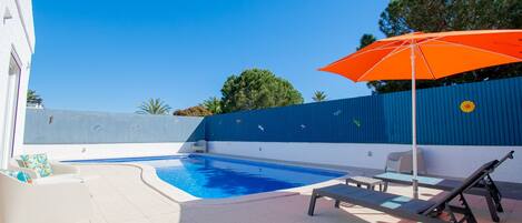 Piscina con sillas de descanso y una sombrilla, rodeada por un muro azul y árboles.