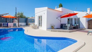 Una piscina con un edificio blanco y sombrillas naranjas al fondo.
