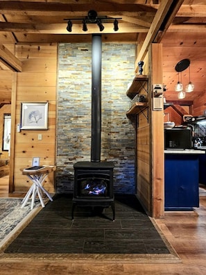 A cozy room with a stone wall, a black wood-burning stove, wooden beams, and a small table with a chair. - Romantic Creekside Cabin Retreat with Hot Tub, Fire Pit and Peaceful Forest Views in Sautee Nacoochee, Georgia (Sautee Nacoochee)