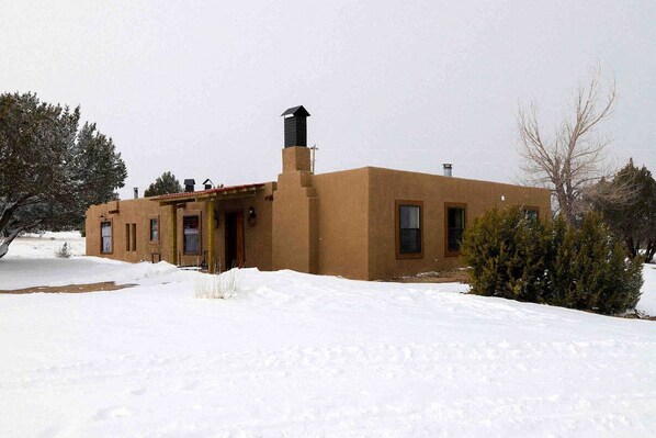Una edificación de un piso con chimenea y un pequeño porche, sobre un paisaje cubierto de nieve.