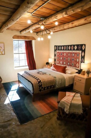 Un dormitorio con techo de madera, una cama con cabecera estampada, una mesita de noche con lámpara, un ventanal con cortinas rojas y una pequeña mesa con una planta.