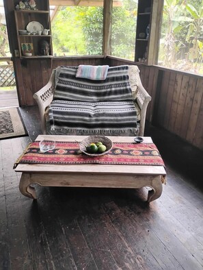 A wooden bench with a woven seat and backrest, a woven table with a bowl of fruit, and a view of greenery through a window.