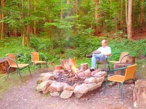 Eine Person sitzt am Lagerfeuer, umgeben von Stühlen, in einer Waldlandschaft.