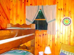 A wooden cabin interior with a bunk bed, a window with curtains, a lamp, and a wall decoration.