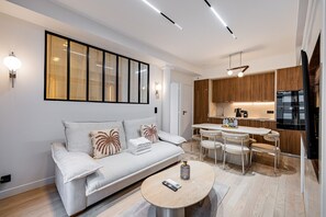 Ein modernes Wohnzimmer mit einer Couch, einem Esstisch mit Stühlen, einer Küchenzeile mit Holdekor und einem großen Fenster mit Jalousien.