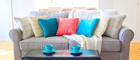 Un sofá gris con varios cojines, un reposapiés negro y dos tazas color turquesa encima.