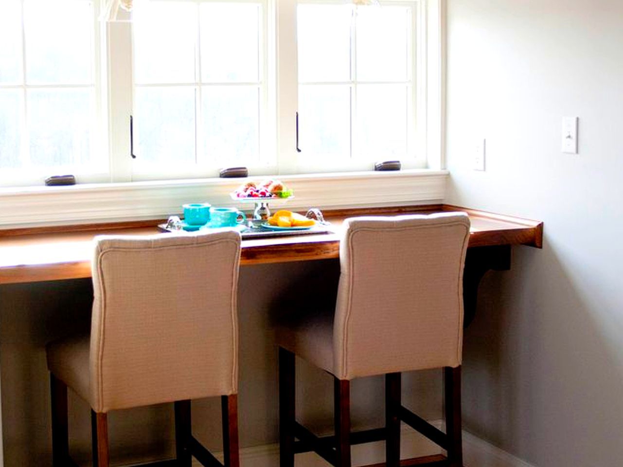 Un comedor con una mesa de madera y dos sillas, una ventana con persianas, y una pequeña mesa con comida y bebidas.