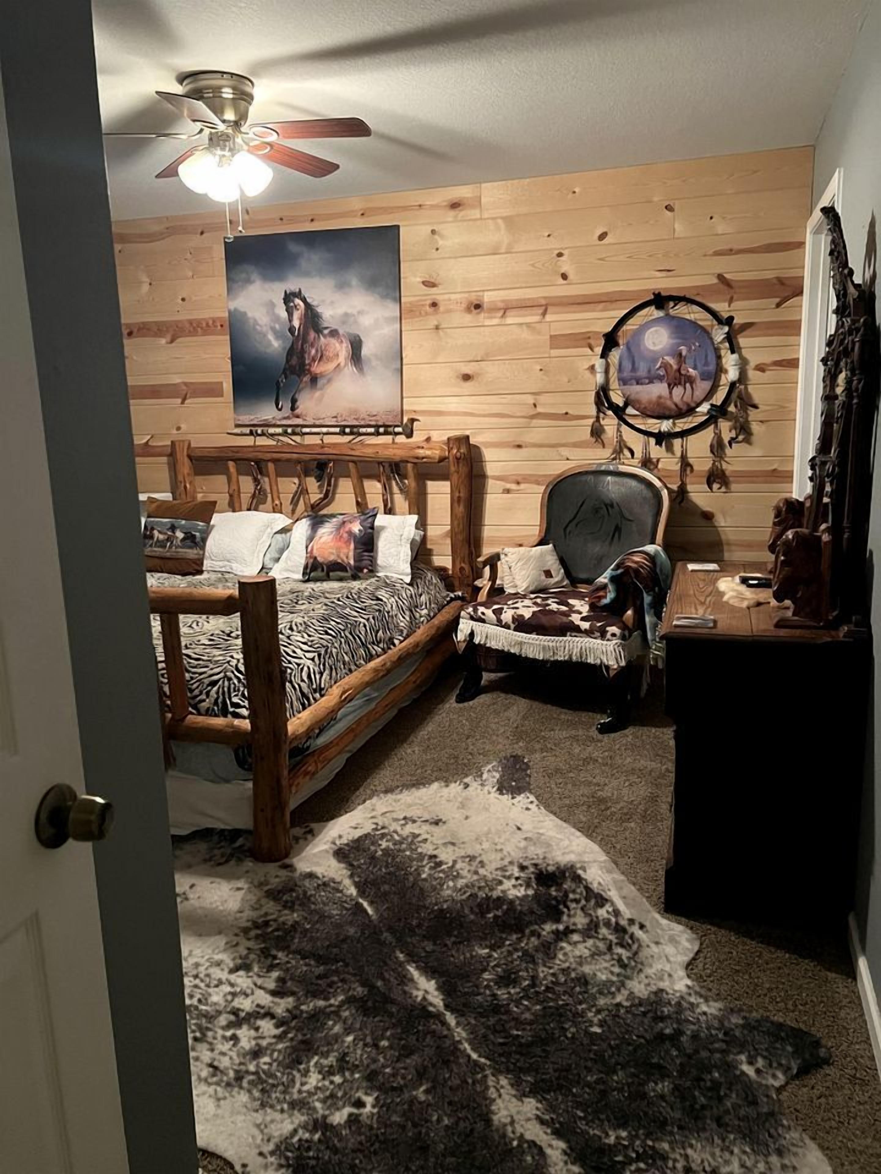 A bedroom with a wooden bed, a chair, a desk, a ceiling fan, a wall painting, and a dreamcatcher.