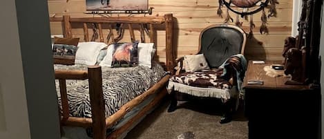 Un dormitorio con una cama de madera, una silla, un escritorio, un ventilador de techo, una pintura en la pared y un atrapasueños.
