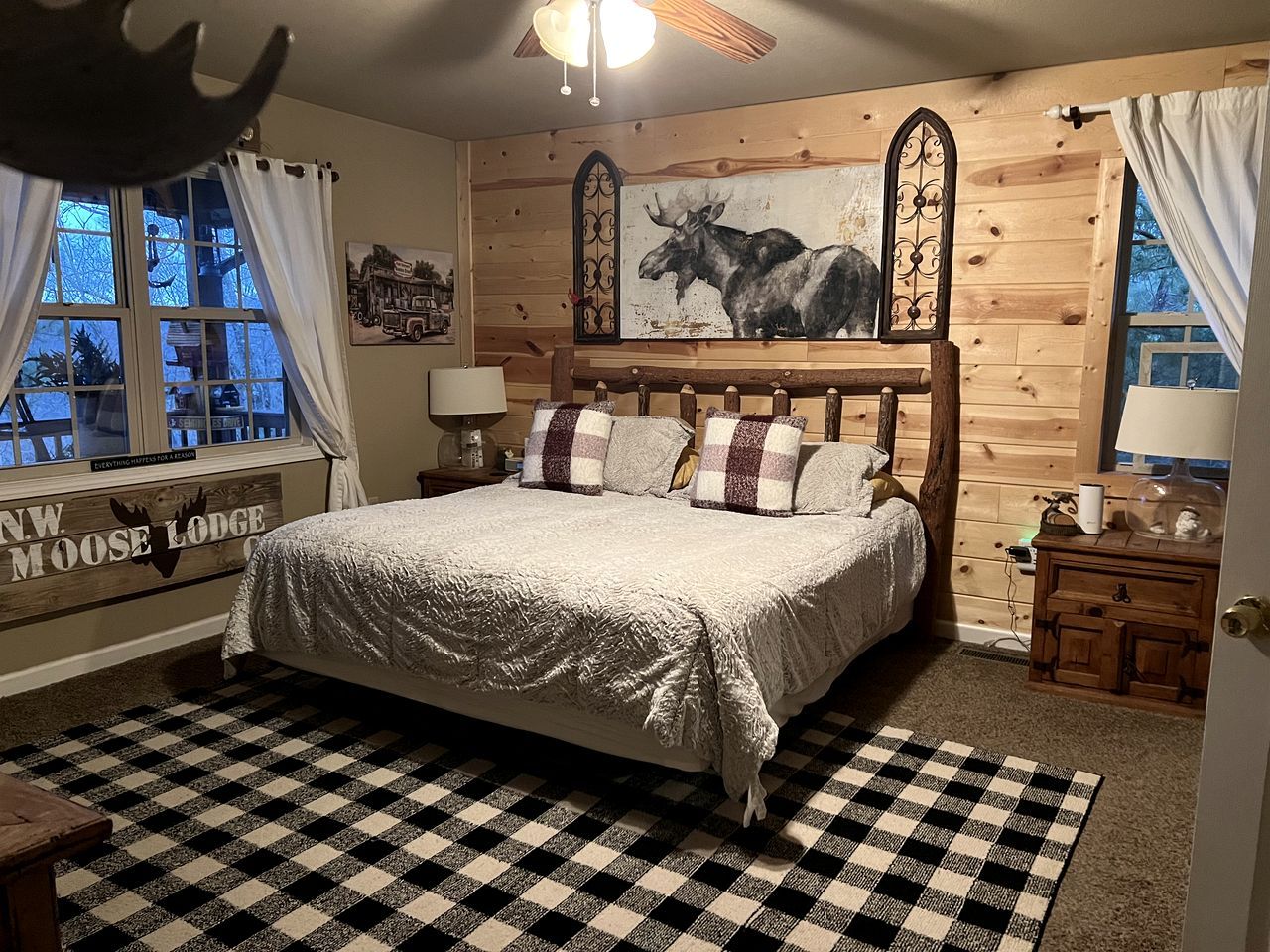 A cozy room with a large bed, a checkered rug, wooden walls, a moose painting, and a window with curtains.