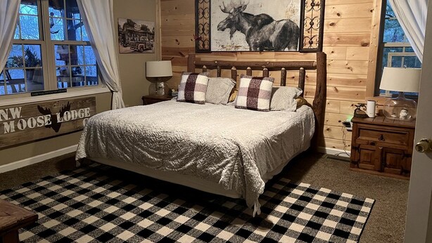 A cozy room with a large bed, a checkered rug, wooden walls, a moose painting, and a window with curtains. - Spacious Camping Cabin Perfect for Families in the Blue Ridge Mountains of Georgia (Blairsville)