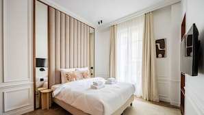 Ein Hotelzimmer mit einem großen Bett, einem Nachttisch, einem Spiegel, einem Flachbildfernseher und einem Fenster mit Vorhängen.