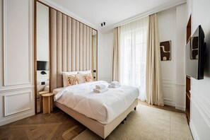 Ein Hotelzimmer mit einem großen Bett, einem Nachttisch, einem Spiegel, einem Flachbildfernseher und einem Fenster mit Vorhängen.