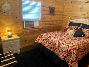A wooden-paneled room with a bed, a nightstand, a lamp, and a window with blinds. - Rustic Dog-Friendly Cabin with Private Deck and Hot-Tub in Texas (Wimberley)
