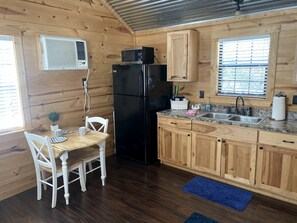 A wooden cabin-style kitchen with a black refrigerator, wooden cabinets, a granite countertop, and a dining table with white chairs. - Rustic Dog-Friendly Cabin with Private Deck and Hot-Tub in Texas (Wimberley)