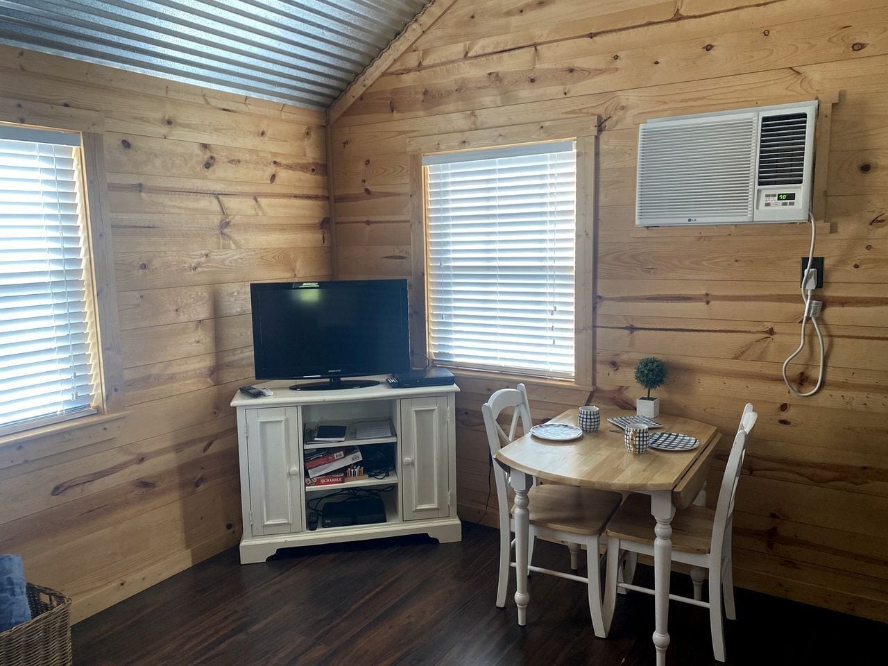 A wooden cabin interior with a television on a cabinet, a dining table with chairs, and a window with blinds.