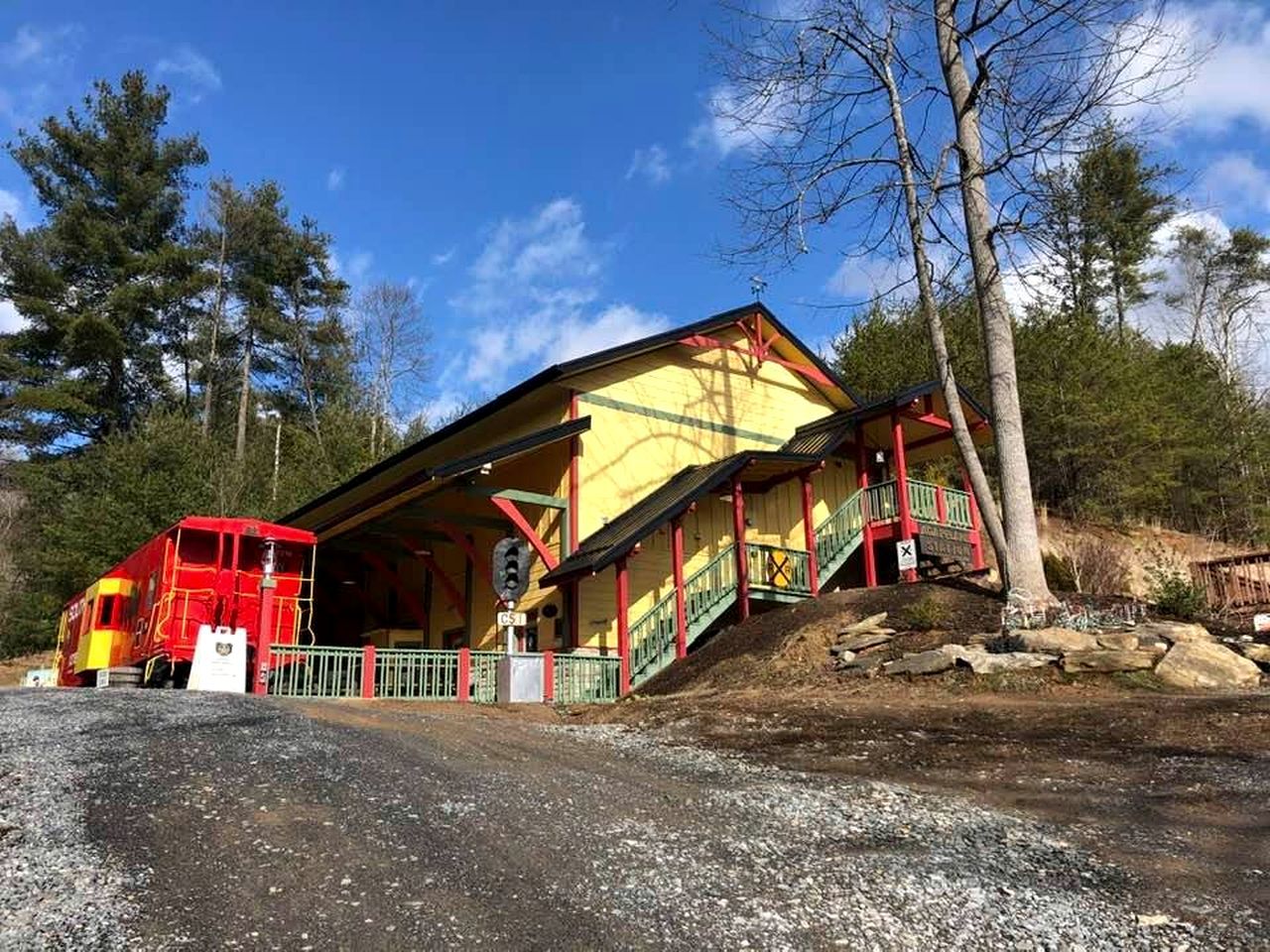 Un edificio con fachada amarilla y detalles rojos, una escalera que lleva a un área cubierta, y un vagón de tren rojo estacionado al frente.