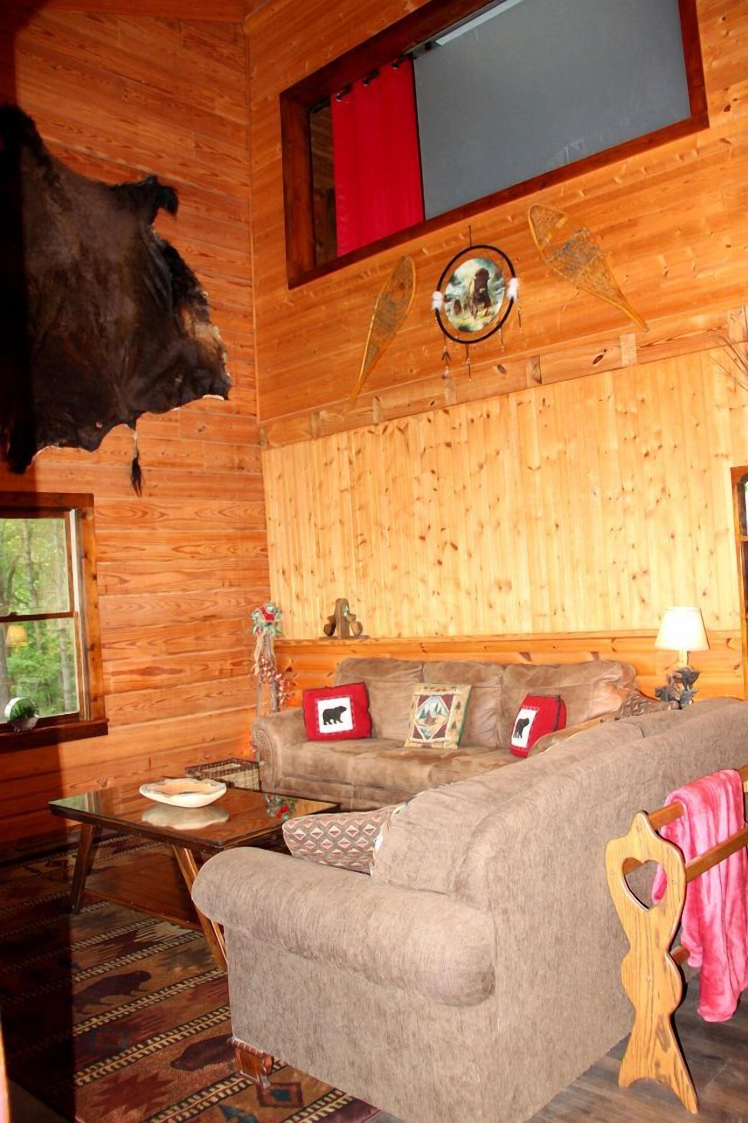 A log cabin interior with a sofa, a mounted animal head, and a window with a view of trees.