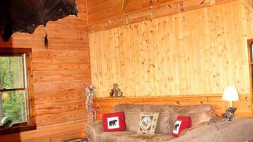 A log cabin interior with a sofa, a mounted animal head, and a window with a view of trees.