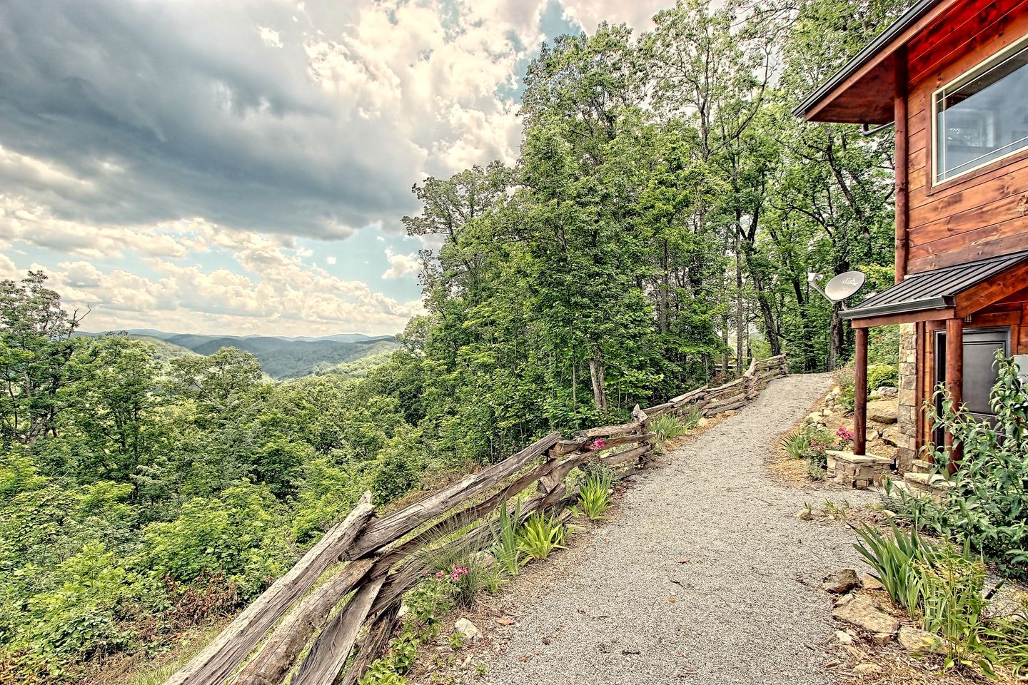A wooden cabin with a gravel path leading to it, surrounded by lush greenery and mountains in the background.