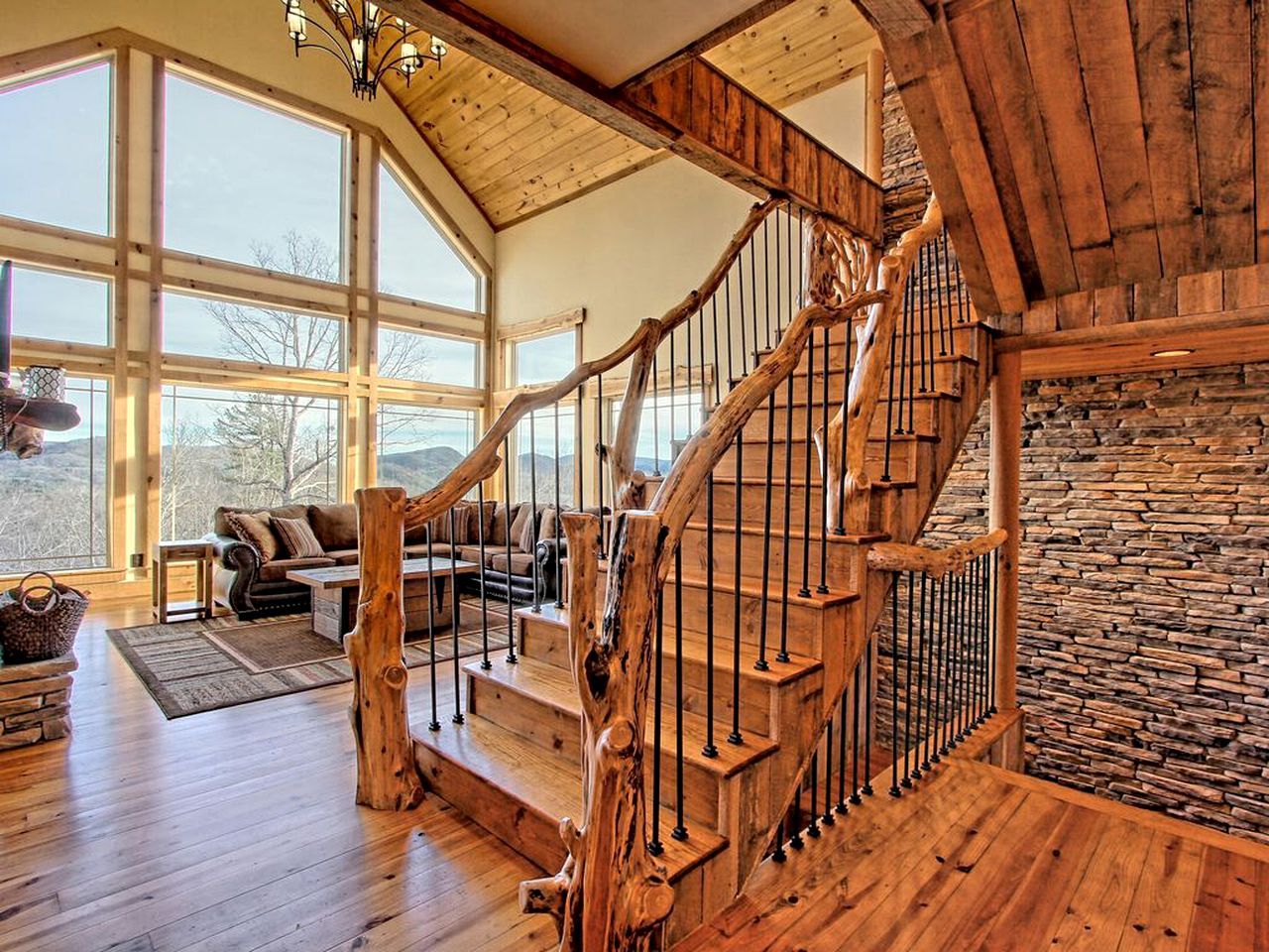 A wooden staircase with a stone wall, a living room with a sofa and a fireplace, and large windows.