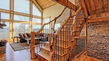 A wooden staircase with a stone wall, a living room with a sofa and a fireplace, and large windows.