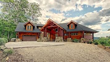 A large, rustic log cabin with a stone foundation, a central porch, and a gravel driveway.