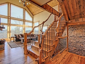 A wooden staircase with a stone wall, a living room with a sofa and a fireplace, and large windows.