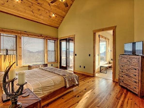 Une chambre avec un plafond en bois, un lit recouvert d’une couvre-lit à motifs, une commode et une vue à travers une porte.