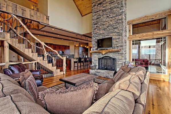 A spacious living area with a stone fireplace, a wooden staircase, and a large window overlooking a scenic view.