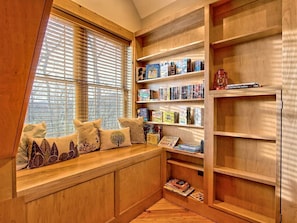 A cozy reading nook with a built-in bench, wooden shelves, and a window with blinds.