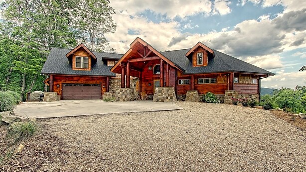 A large, rustic log cabin with a stone foundation, a central porch, and a gravel driveway.