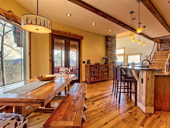 A rustic interior with a wooden dining table, benches, a stone fireplace, and a kitchen area with a bar.