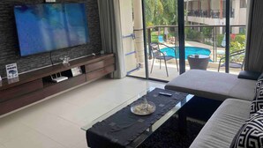 A living room with a TV, a sofa, a glass coffee table, and a view of a pool and outdoor seating. - Apartment Eliza Samui in Garden Resort With Shared Pool (Koh Samui)