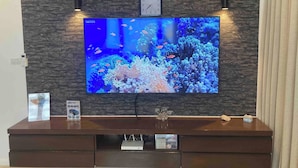 A television mounted on a stone wall, a clock above it, and a wooden entertainment center below. - Apartment Eliza Samui in Garden Resort With Shared Pool (Koh Samui)
